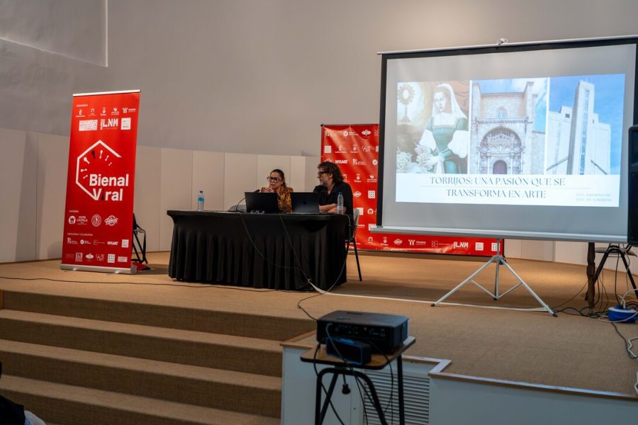 Bienal viral - IP | Slow Radio Two presenters sit at a table on a stage with a projector screen showing a slide about art and transformation, at a Bienal event.